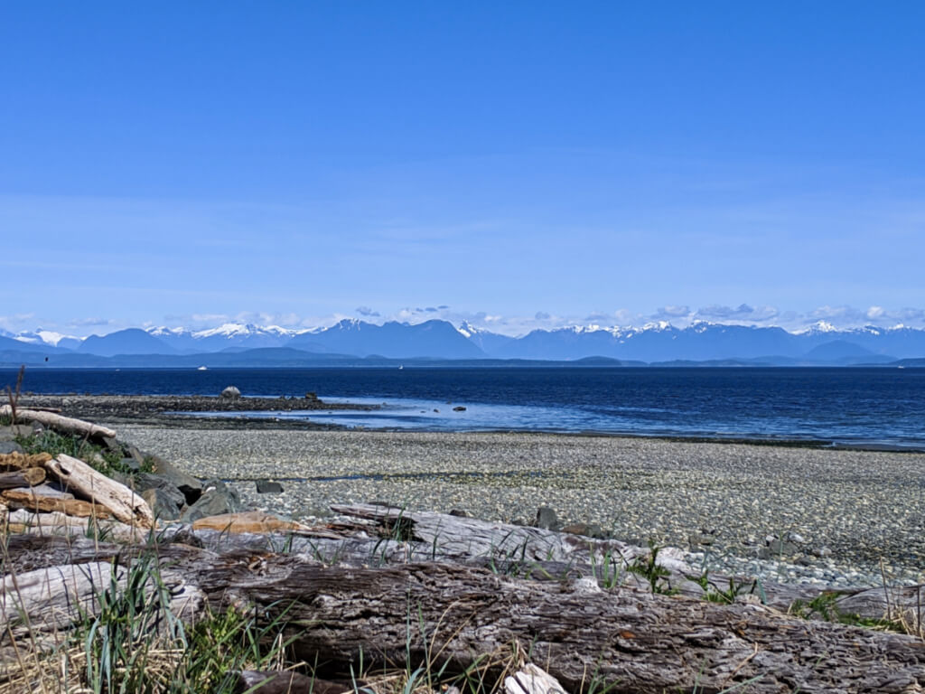 Driftwood lined rocky beach with calm ocean and backdrop of snow capped mountains