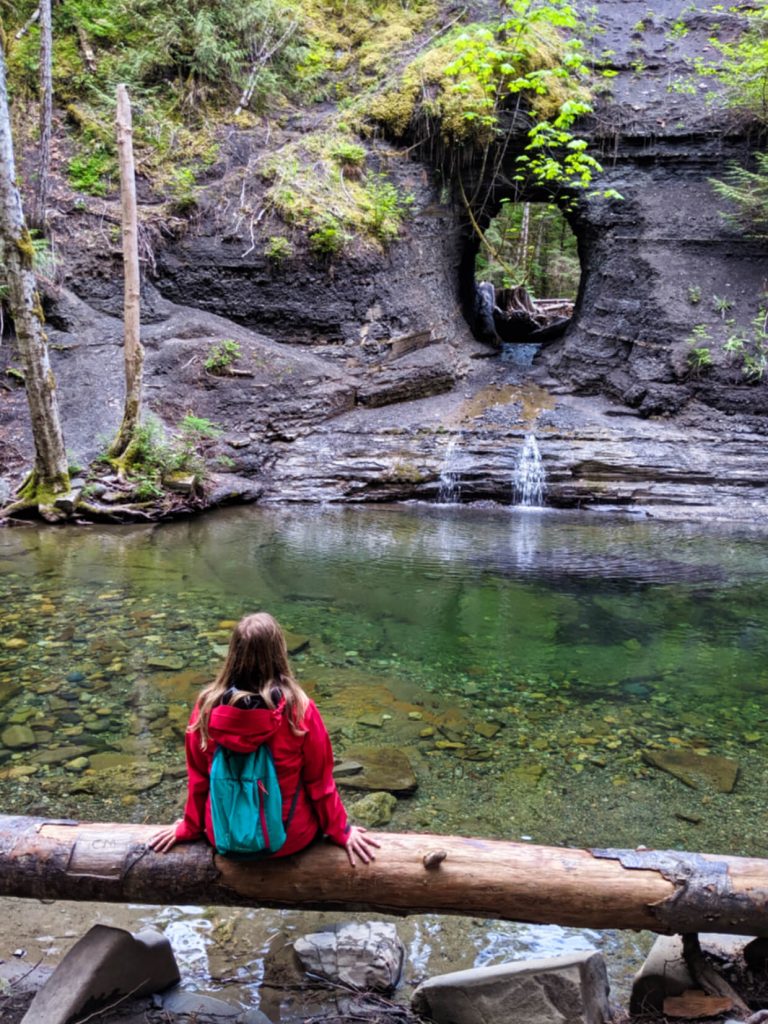 Gemma sat on log looking at thin waterfall dropping through round hole and into turquoise coloured pool