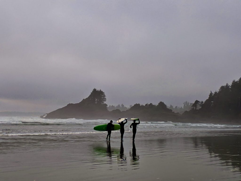 Surfers waling along wet beach in Tofino with boards on hea