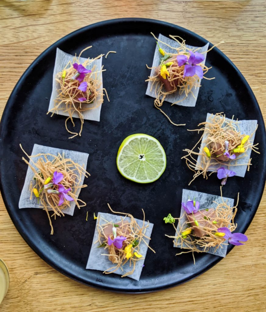 Overhead shot of black plate with small pieces of tuna, dressed with flowers and noodles