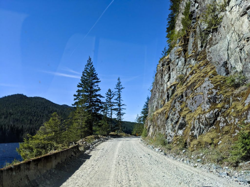 Windshield view of gravel road on Vancouver Island with mossy cliff on right and lake (with barrier) on left